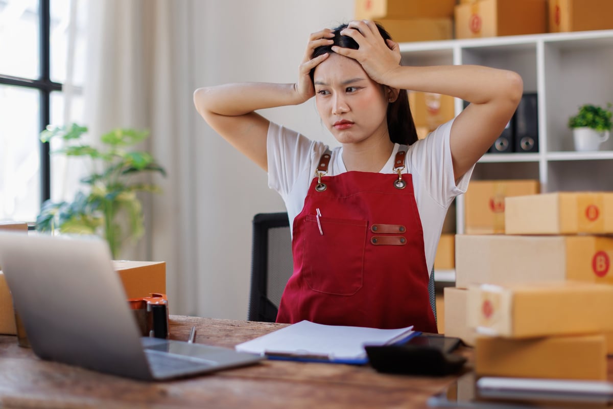 Young adult woman feeling overwhelmed and frustrated, holding her head in hands while working on a laptop at a desk surrounded by shipping boxes for her e-commerce business