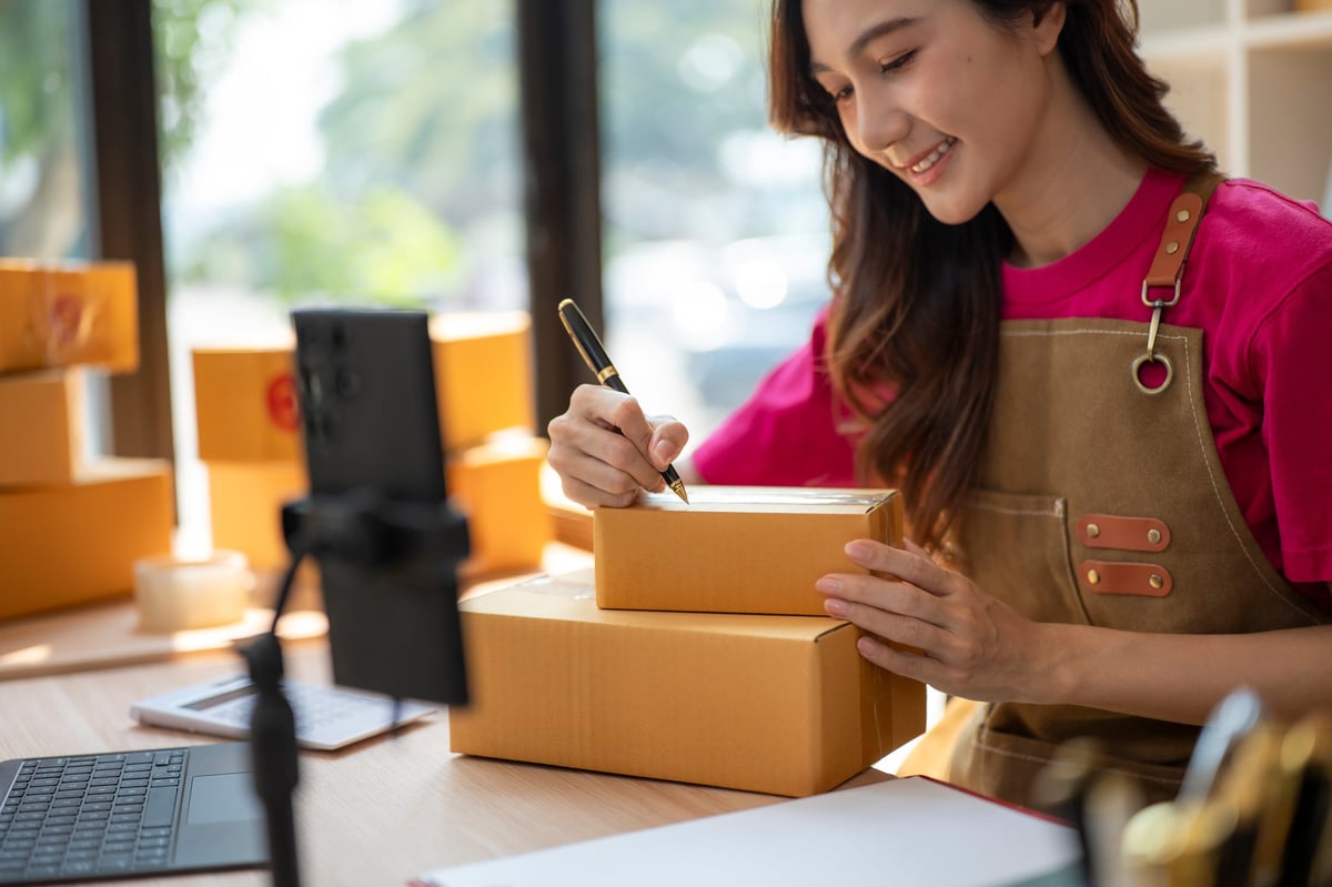 A young woman happily packages orders at her home office desk, showcasing her commitment to customer service and creativity in a vibrant workspace filled with boxes.